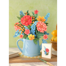 Load image into Gallery viewer, Colorful flowers in a blue watering can on a wooden surface with a green background
